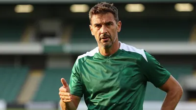 A close-up, waist-up shot of a mid-30s, ruggedly-built, Shamrock Rovers coach, dressed in a plain, dark green and white...